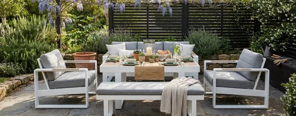 Modern outdoor seating with white frames and grey cushions in a lush garden.